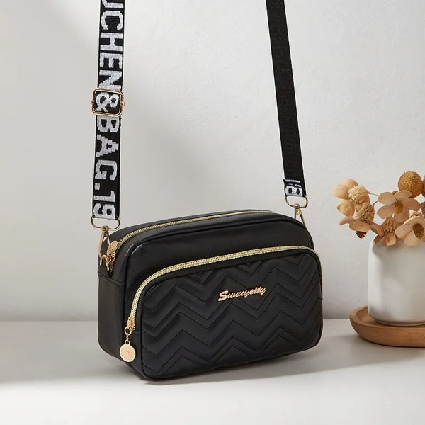 Black quilted crossbody camera bag with a gold letter strap resting on a white surface.