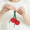 A hand holds a red cherry fruit shape coin purse keychain with gold hardware and green thread.