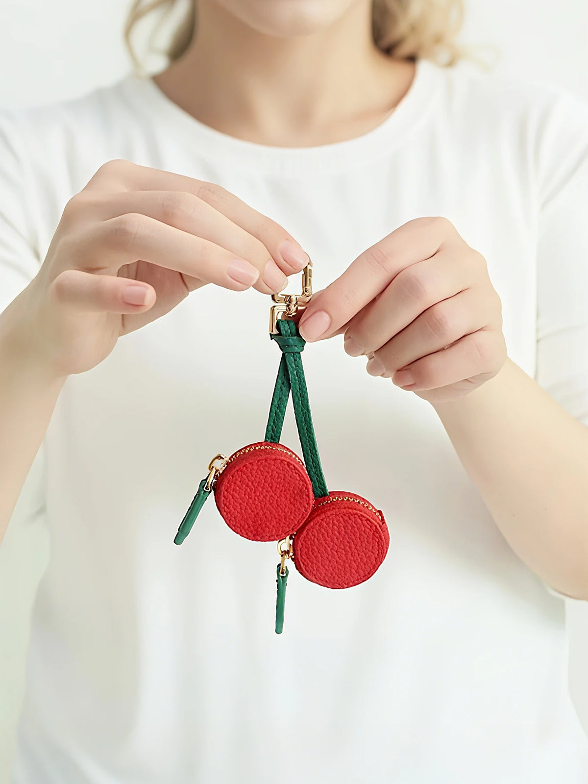 A hand holds a red cherry fruit shape coin purse keychain with gold hardware and green thread.