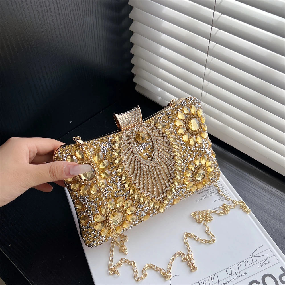 A hand holds a gold rhinestone evening clutch bag against a dark background with white blinds.