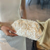 A hand holds a white beaded chain shoulder clutch bag with gold sequins against a light-colored dress.