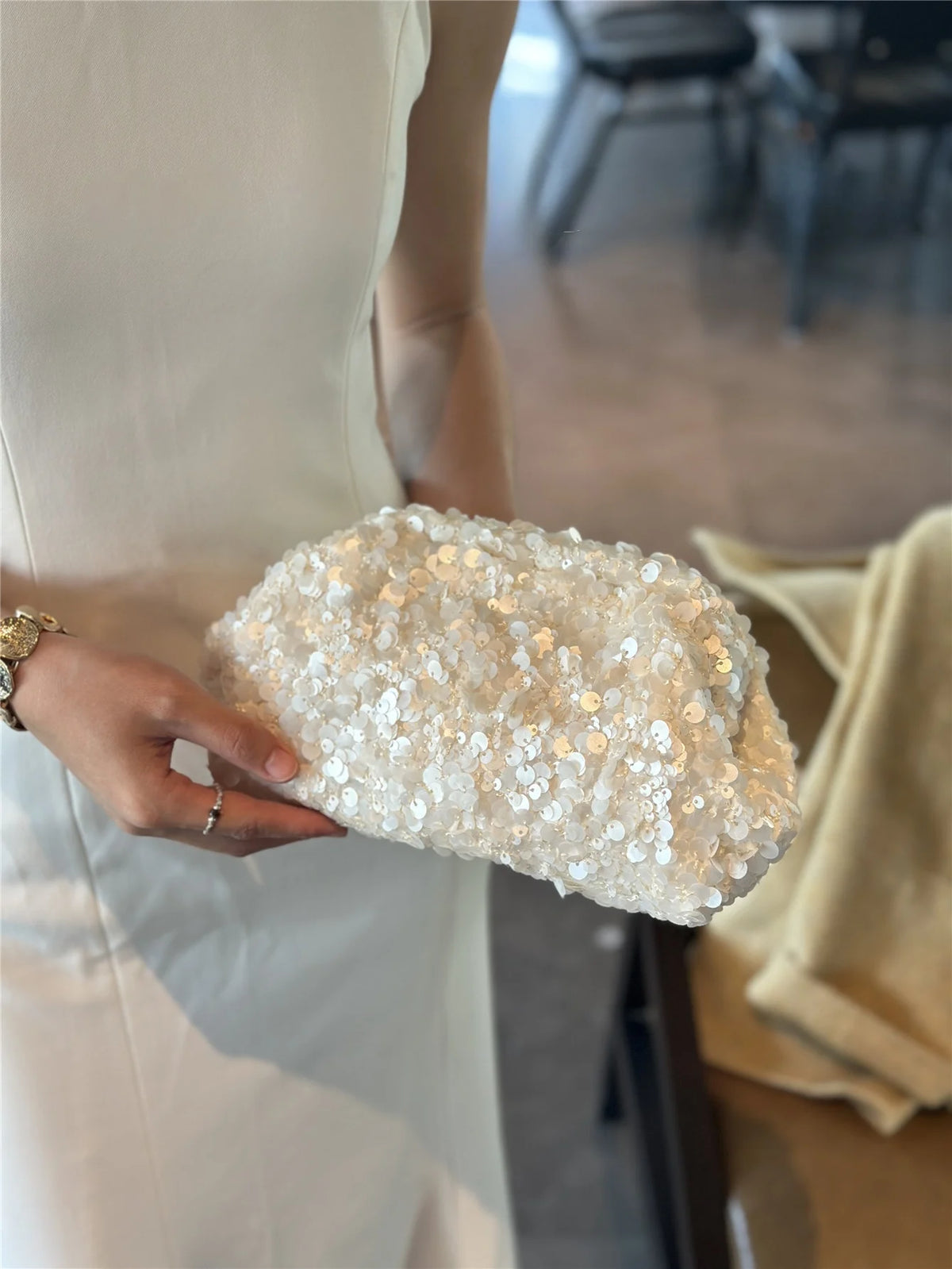 A hand holds a white beaded chain shoulder clutch bag with gold sequins against a light-colored dress.