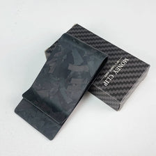 A black carbon fiber money clip is displayed inside its box with a textured pattern.