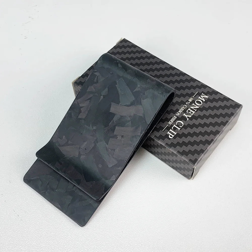 A black carbon fiber money clip is displayed inside its box with a textured pattern.