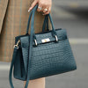 A dark teal genuine leather shoulder bag with gold hardware is held by a person wearing a plaid coat.