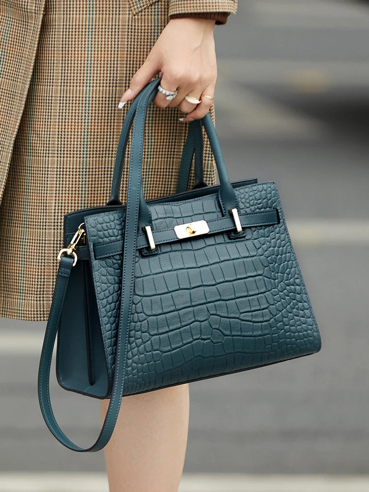 A dark teal genuine leather shoulder bag with gold hardware is held by a person wearing a plaid coat.
