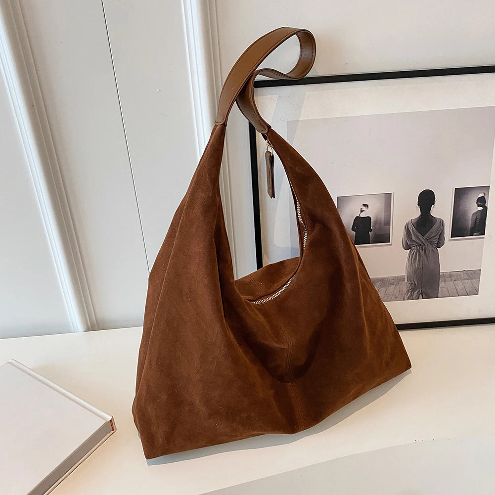 A brown suede slouchy shoulder bag is displayed on a white surface next to a framed photograph.