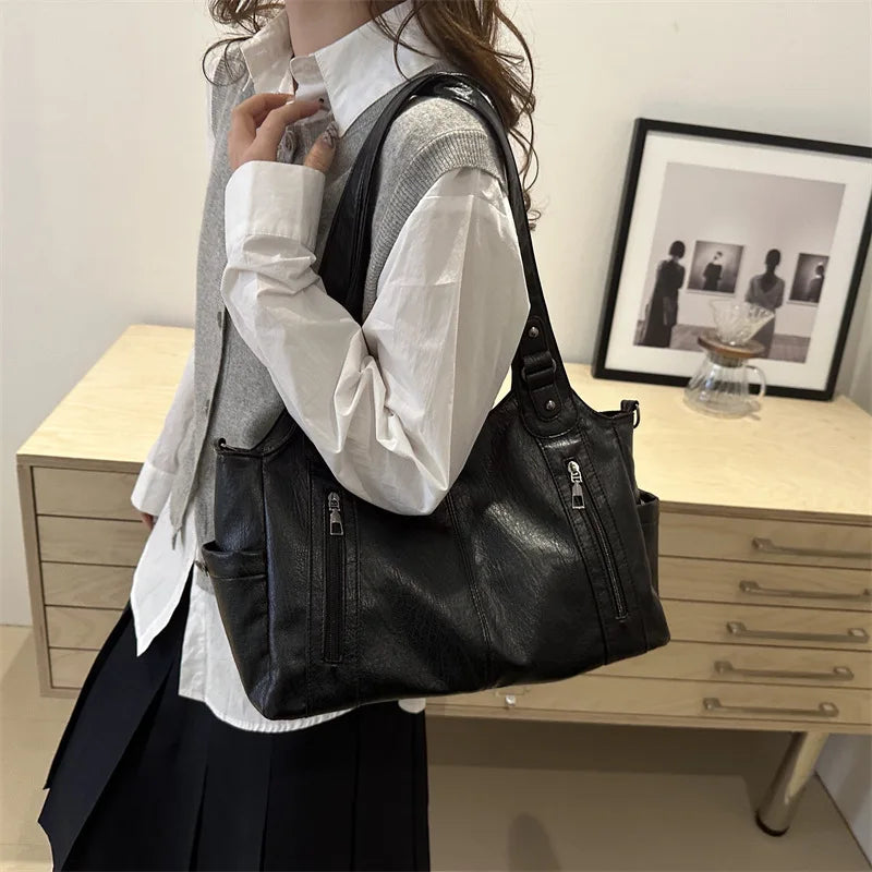 A woman holds a black leather everyday large-capacity shoulder tote bag against a light wooden cabinet.