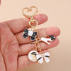 A gold enamel heart keychain with a black bow and white butterfly charm is held in a hand.