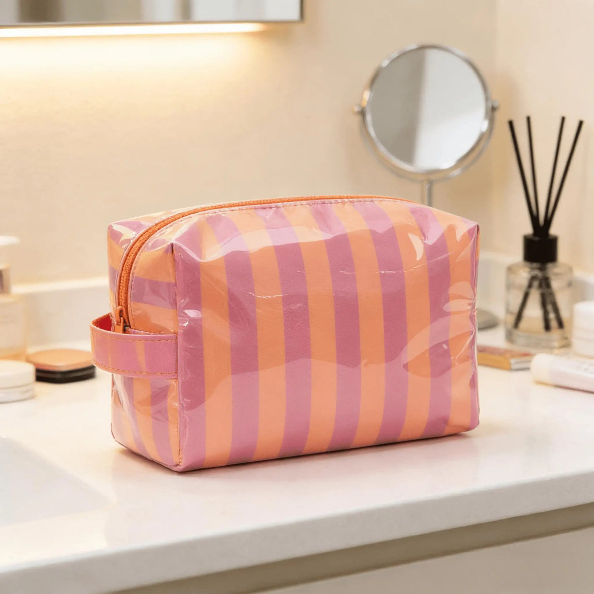 Sac de rangement cosmétique rayé rose et orange posé sur un comptoir blanc à côté d'un miroir.
