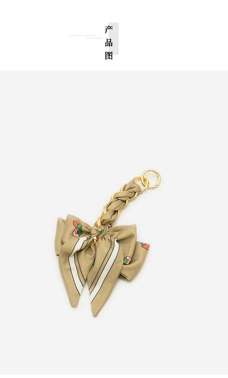 A beige fabric bow bag with a gold chain strap extension and small fruit embellishments.