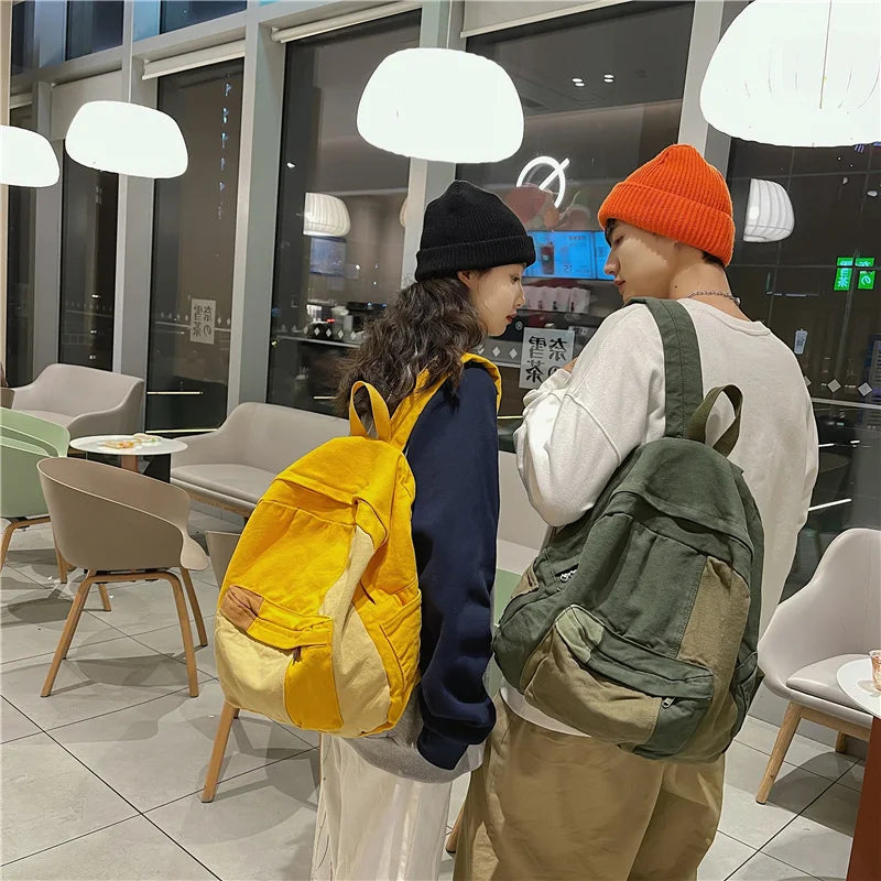 Two students wearing orange and black knit hats are standing in a modern cafe, one carrying a yellow canvas backpack.