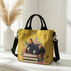 Gelige Tasche mit Blumen-Katzenmuster und schwarzem Riemen auf weißem Untergrund.