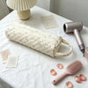 A quilted beige storage bag for a hair dryer and personal items rests on a white tablecloth with a pink comb nearby.