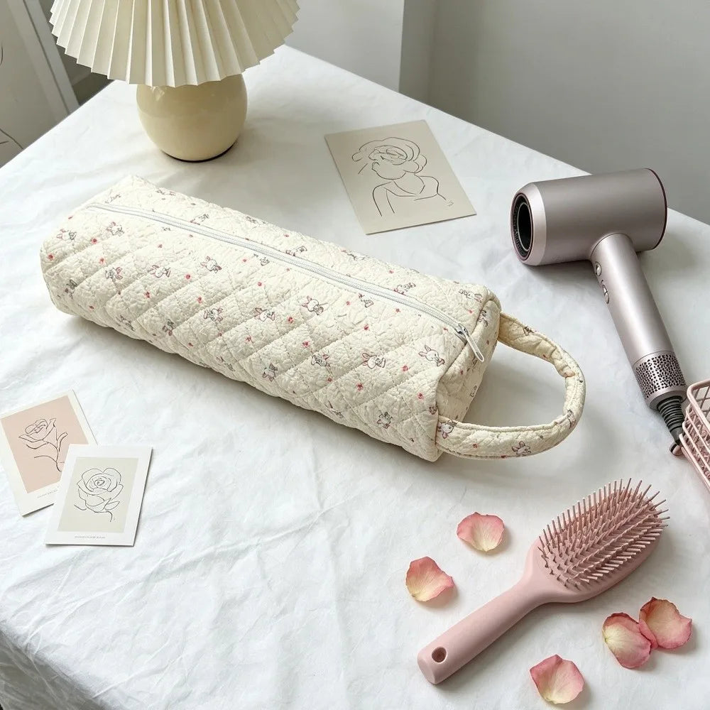 A quilted beige storage bag for a hair dryer and personal items rests on a white tablecloth with a pink comb nearby.