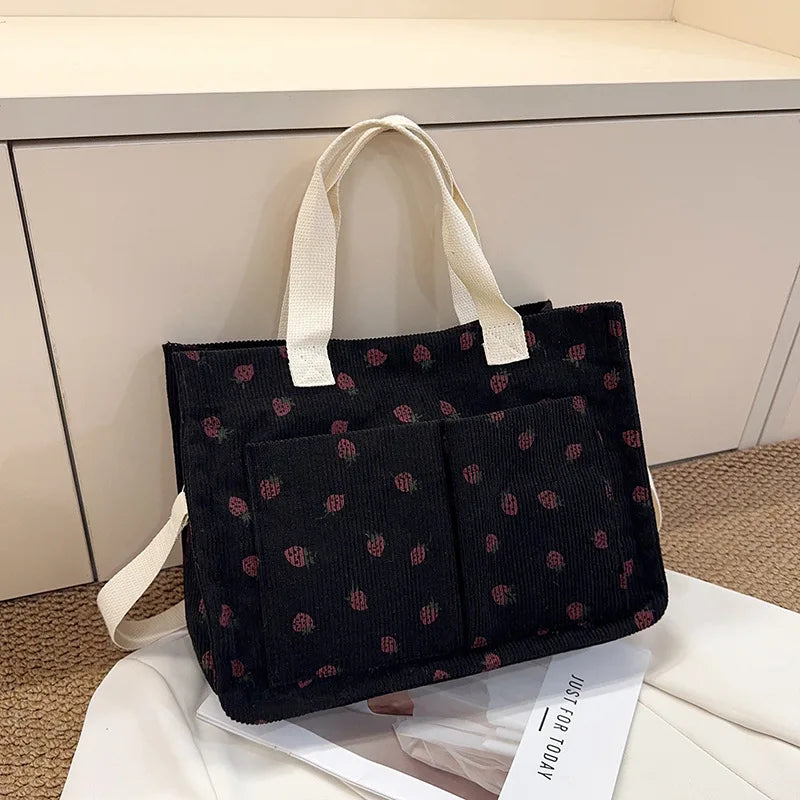 A black canvas tote bag with a repeating strawberry pattern is displayed on a white surface.