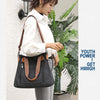 A woman holds a black canvas tote bag with brown leather straps against a white wall.