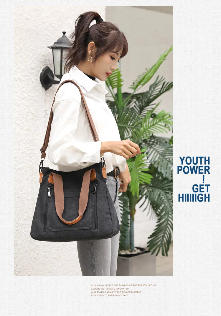 A woman holds a black canvas tote bag with brown leather straps against a white wall.