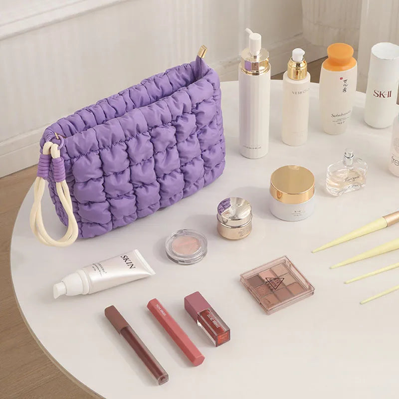 A purple quilted puffer makeup bag is displayed on a white table with various cosmetic products.