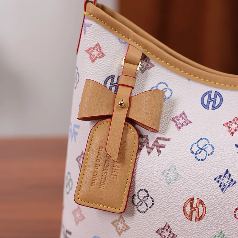 A light pink canvas tote bag with a tan leather strap and bow accent featuring a repeating letter print pattern.