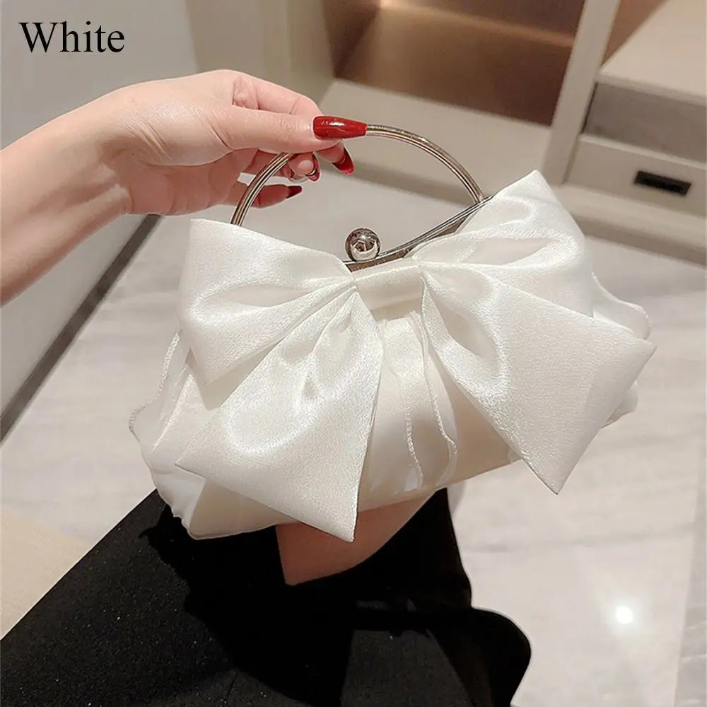 Una mano sostiene un bolso de mano de satén blanco con lazos y una asa dorada.