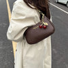 A brown leather shoulder bag with a gold chain and cherry pendants is carried by a person wearing a light beige coat on an as