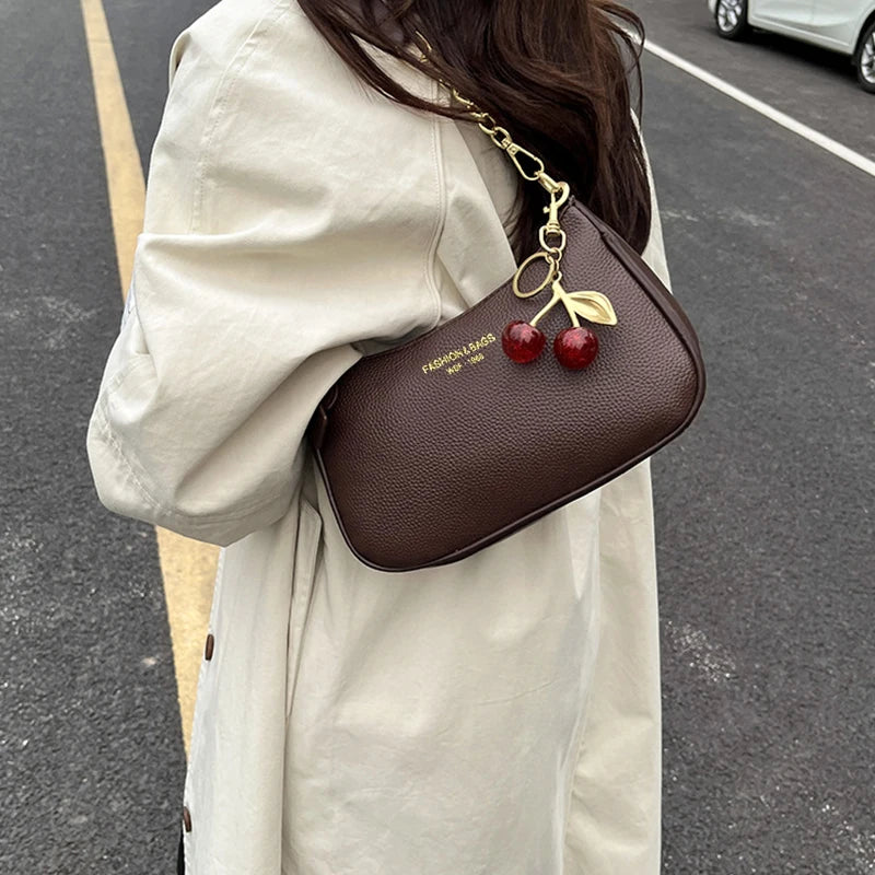 A brown leather shoulder bag with a gold chain and cherry pendants is carried by a person wearing a light beige coat on an as