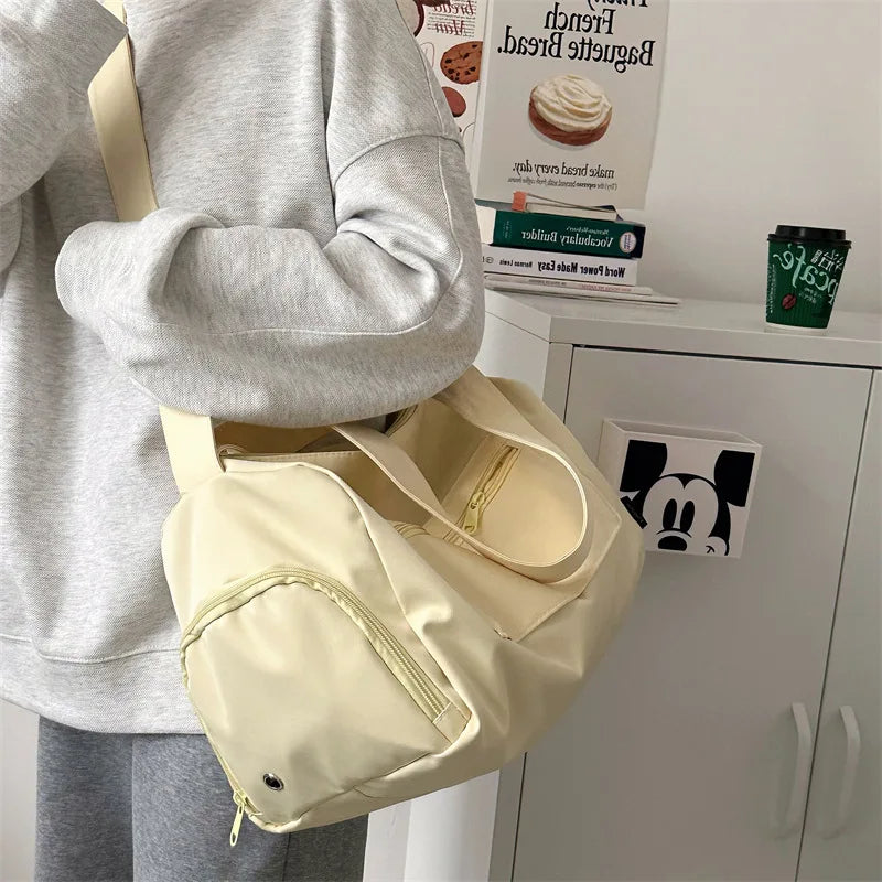 A beige canvas crossbody travel tote bag is shown being held by a person, next to some packaged goods.