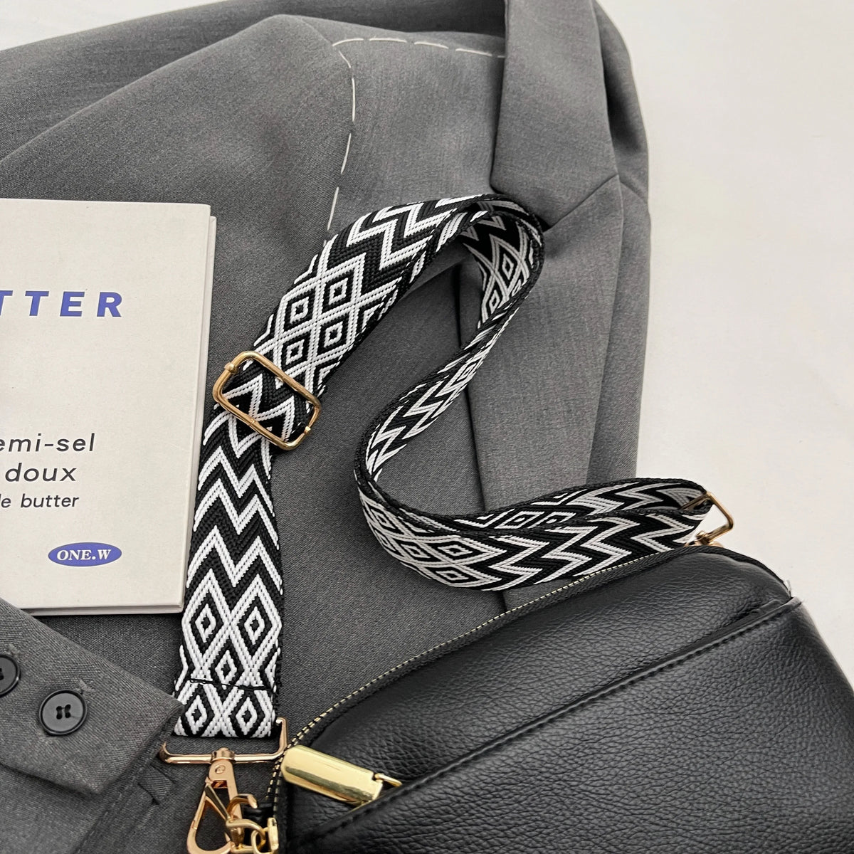A gray jacket features a black and white geometric patterned strap attached to a leather crossbody bag.