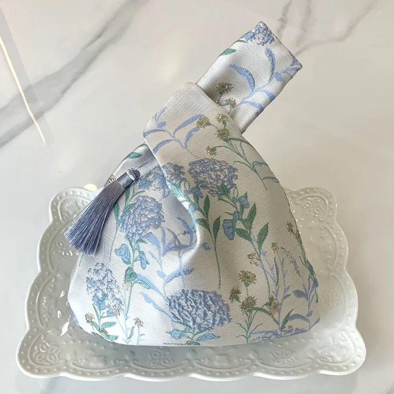 A light blue and green embroidered silk evening clutch with a tassel resting on a white decorative plate.