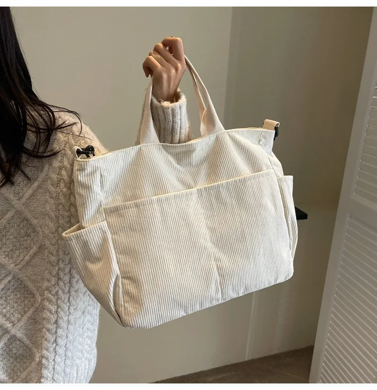 A white corduroy multi-pocket tote bag is hanging on a door, held by a person's hand.