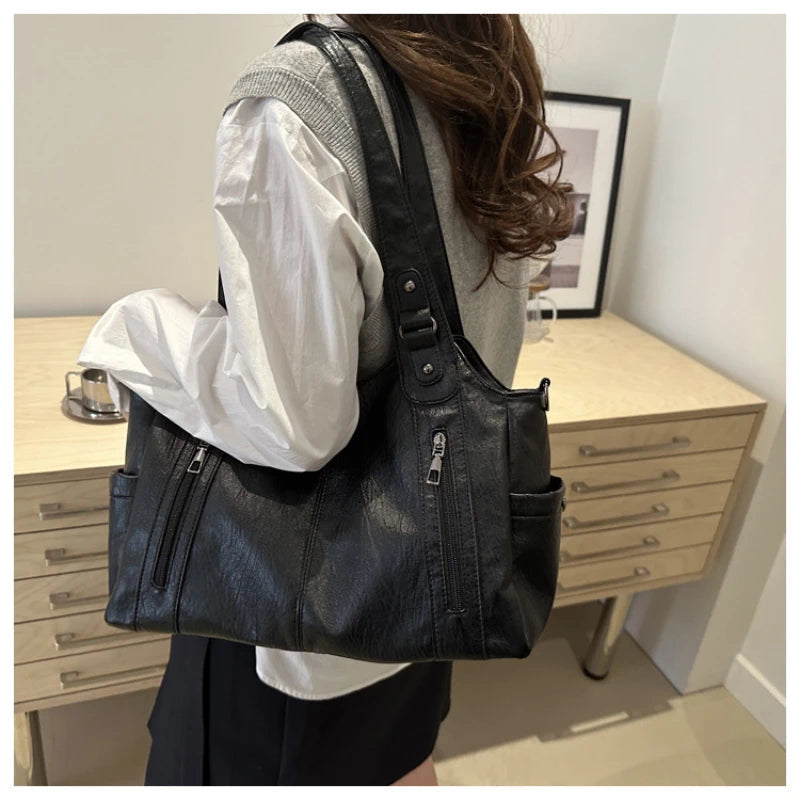 A person is holding a black leather everyday large-capacity shoulder tote bag in front of a wooden dresser.
