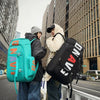Two people stand outdoors carrying black and teal backpacks on a city street.