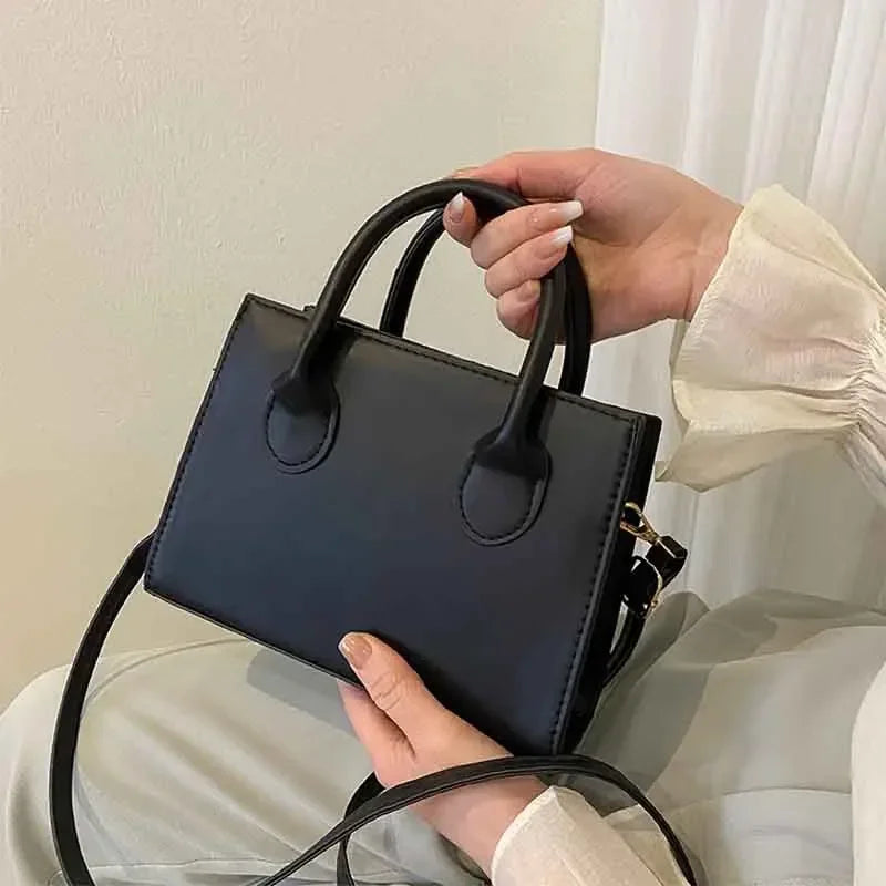 Une main tient un sac à bandoulière noir en cuir PU minimaliste sur fond blanc.