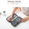 A hand is opening a dark gray multi-pocket travel document organizer filled with various currency and documents on a white ta