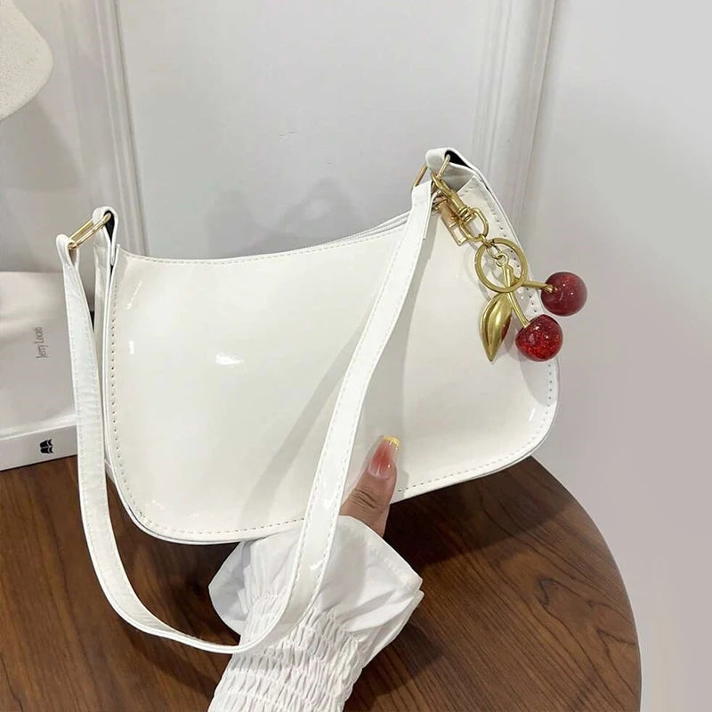 A white PU leather shoulder bag with a gold chain and a red cherry pendant is displayed on a wooden surface.