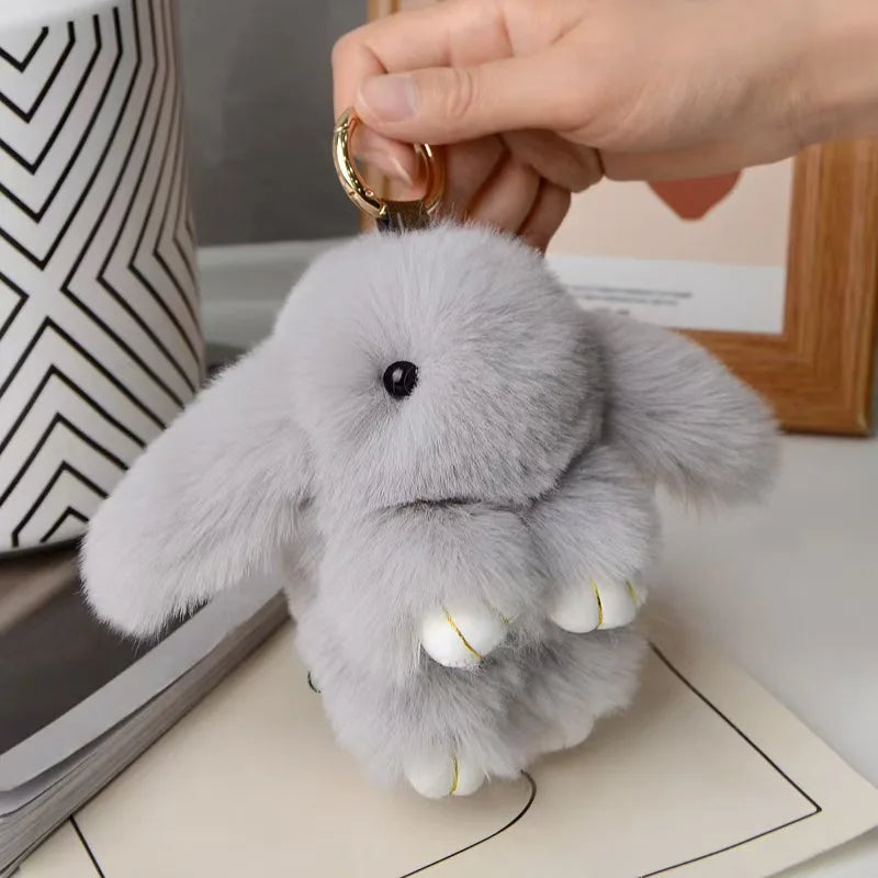 A soft gray plush rabbit charm is being held by a hand, positioned on a light-colored surface.