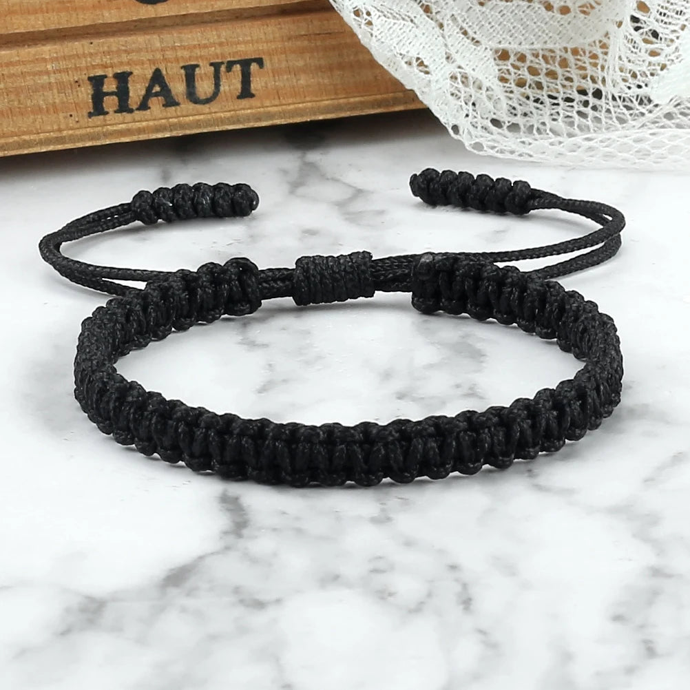 A black braided rope bracelet is displayed on a white marble surface next to a wooden box.