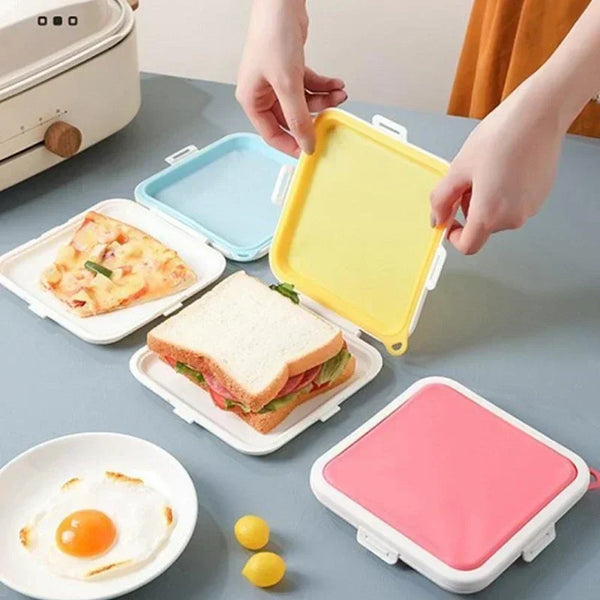 Hands are placing a yellow silicone reusable sandwich and lunch container onto a table next to a plate of food.