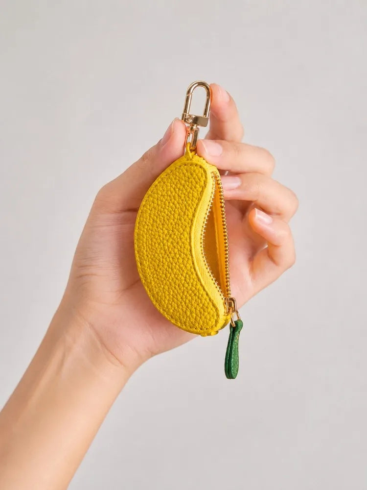 A hand holds a yellow fruit-shaped PU leather coin purse keychain with a gold clasp and green tassel.