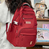 A red lightweight nylon college backpack is shown from the back, resting on a wooden shelf next to a stuffed animal.
