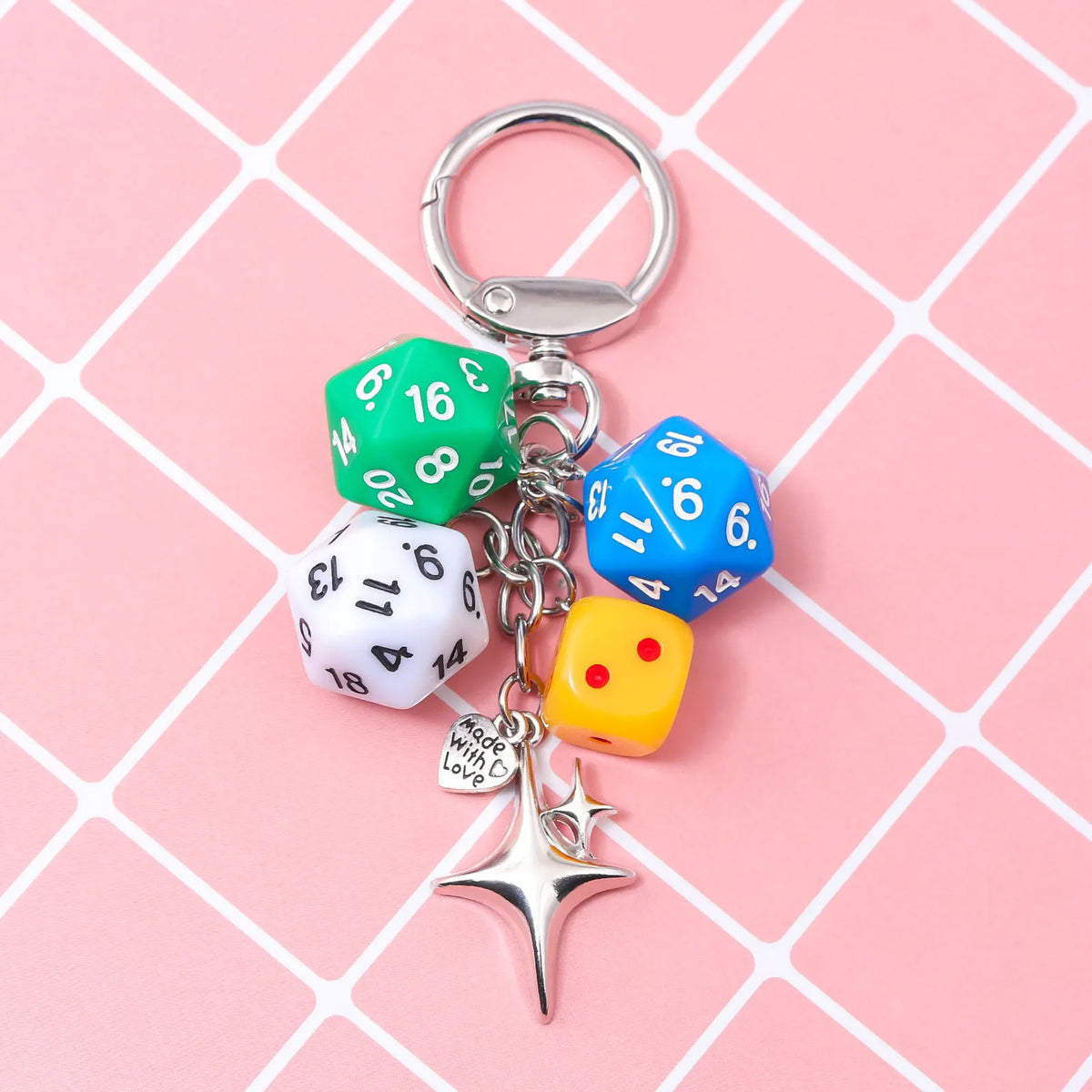 A silver keychain charm featuring a green and blue dice, a yellow dice, and a star charm on a pink grid background.