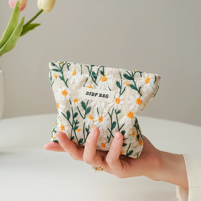 A hand holds a floral canvas makeup bag featuring white and green botanical embroidery.
