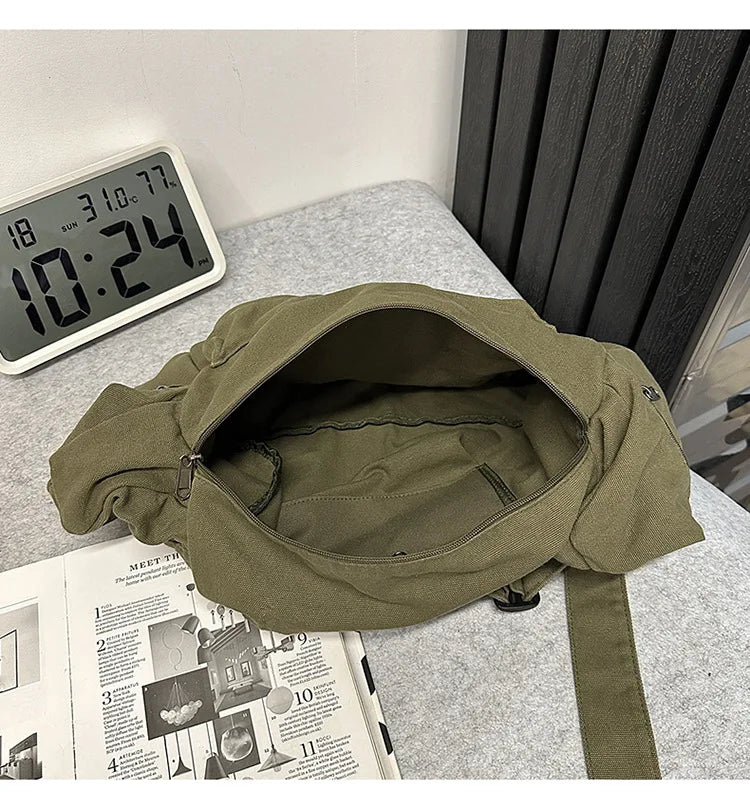 A olive green cotton canvas shoulder crossbody bag lying on a gray surface next to a digital clock and some papers.