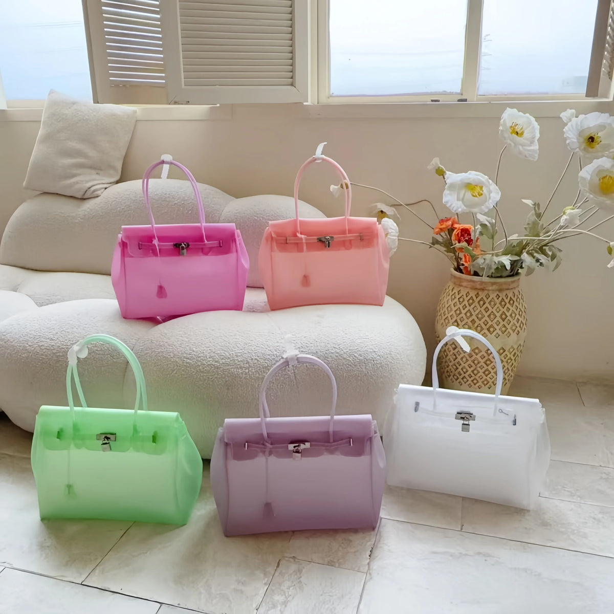 Eine Sammlung von fünf pastellfarbenen Sommer-Jelly-Schultertaschen auf einem weißen Sofa am Fenster.