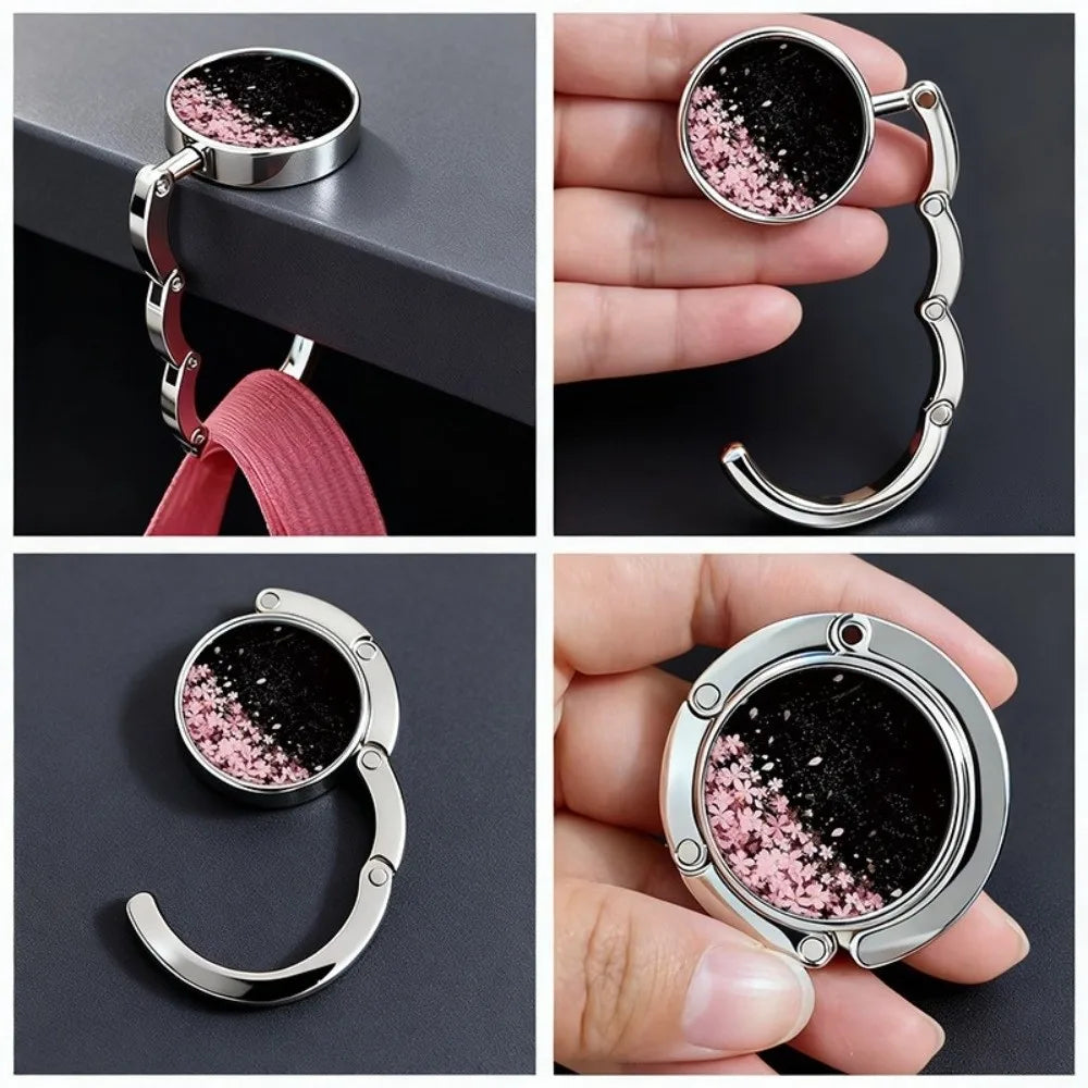 A close-up of a round metal bag hanger with a pink and black glitter finish being held by a hand.
