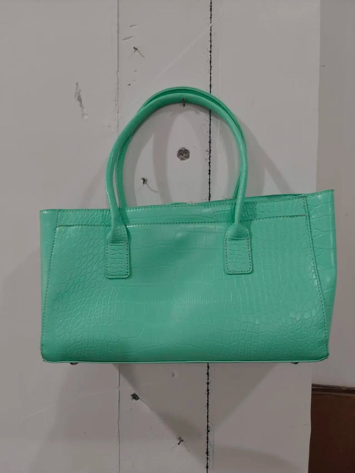 A mint green crocodile embossed leather tote bag with a handle and magnetic closure displayed against a white wooden backgrou