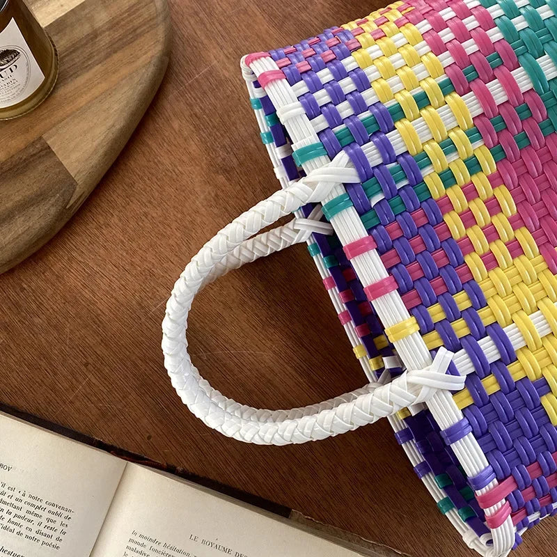 A hand woven PVC vegetable basket tote bag with multicolored stripes and a white handle resting on a wooden surface.