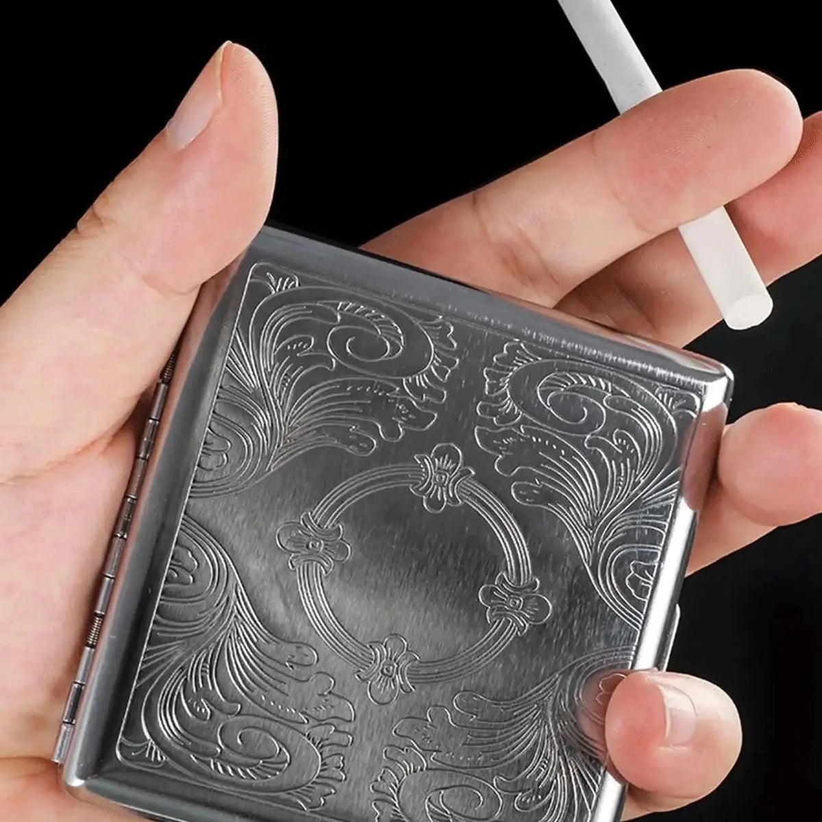 A hand holds a decorative metal cigarette storage box with intricate embossed scrollwork.