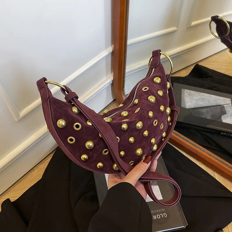 A dark purple faux suede hobo crossbody bag with gold rivet accents being held by a hand.